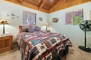 A bedroom with a large bed, wooden ceiling, two windows with blinds, a bedside table with a lamp, and a floor fan.