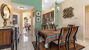 Uma sala de jantar com uma mesa posta para quatro pessoas, rodeada por cadeiras de madeira. Um lustre pendurado sobre a mesa, e um painel decorativo visível na parede.