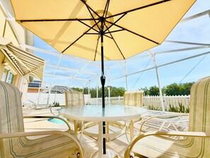A covered outdoor seating area with a glass table and chairs, a pool in the background, and a white picket fence.