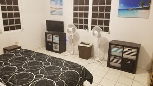 A bedroom with a bed, a TV on a cabinet, two standing fans, a trash bin, and a cabinet with drawers.