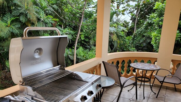 A NEXGRILL barbecue grill with four burners, a side burner, and a chimney, placed on a balcony with a tiled floor and a table with chairs.