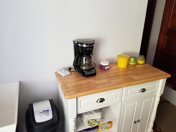 A kitchenette with a coffee maker, a trash bin, and a small cabinet with a towel and a box.