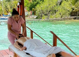 En massagebehandling på ett trädäck vid en turkosfärgad pool, med en tropisk miljö som bakgrund.