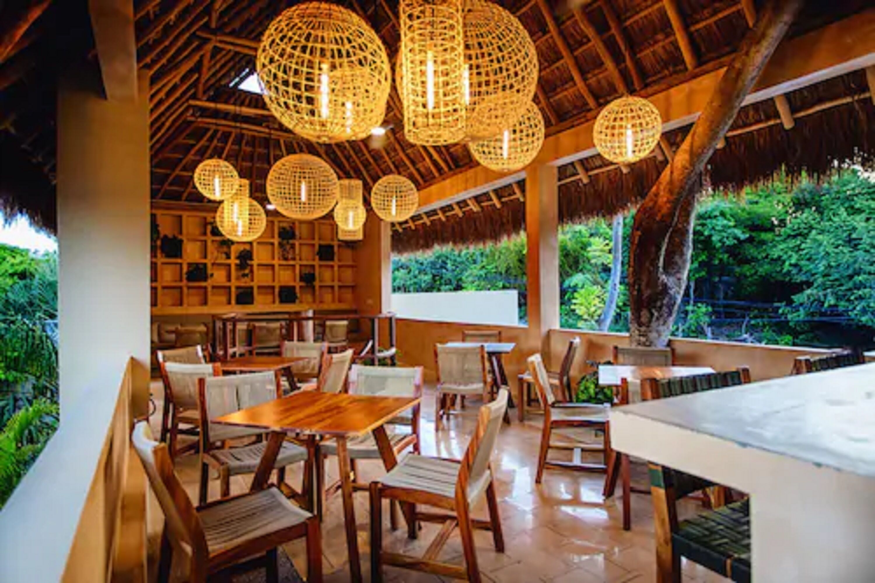 Un área de comedor al aire libre con mesas y sillas de madera, rodeada de exuberante follaje y una estructura de techo de paja.