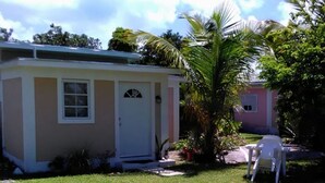 A small, single-story house with a white door and window, surrounded by a well-maintained lawn and a palm tree. - The Colonial Rental Suite, Moss Town Exuma (Moss Town)