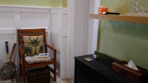 A room with a wooden chair with a cushion, a small table with a plant, and a shelf with decorative items. - The Colonial Rental Suite, Moss Town Exuma (Moss Town)