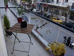 A balcony with a wooden table, potted plants, and a view of a busy street with cars and buildings. - Mini suite neighboring the centre (Thessaloniki)
