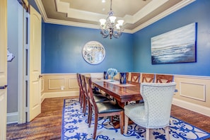 A dining room with a wooden table, chairs, a chandelier, and a blue wall with a painting.