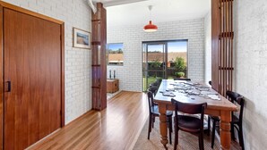 A dining area with a wooden table and chairs, white brick walls, and a view of a garden through large windows. - Fi s Beach House directly opposite beach and park! (Port Macquarie)