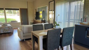A modern living room with a dining area, featuring a sofa, a dining table with chairs, a kitchen area with cabinets, and a large window with blinds. - Fi s Beach House directly opposite beach and park! (Port Macquarie)
