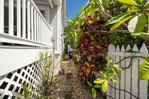 A narrow pathway flanked by white fences and lush greenery, leading to a building with a white exterior.