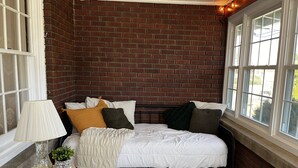 A bedroom with a brick wall, a bed with multiple pillows, a bedside table with a lamp, and a window with curtains.