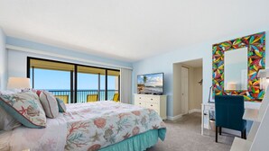 A bedroom with a bed, a TV, a desk, a chair, a mirror, and a balcony with a view. - Ocean Sands 1107: Amazing Gulf Front Retreat (Madeira Beach)