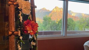 Ein hölzerner Spiegel mit floralen Verzierungen auf einem roten Regal, ein weißer Stuhl und ein Blick auf Berge durch große Fenster.