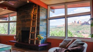 Ein gemütliches Wohnzimmer mit Kamin aus Stein, einer bequemen Couch und großen Fenstern mit Blick auf rote Felsformationen.