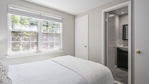 A bedroom with a bed, a window with blinds, a door leading to a bathroom, and a mirror. - BickelScape: Private Yard & Near DC (Sterling)