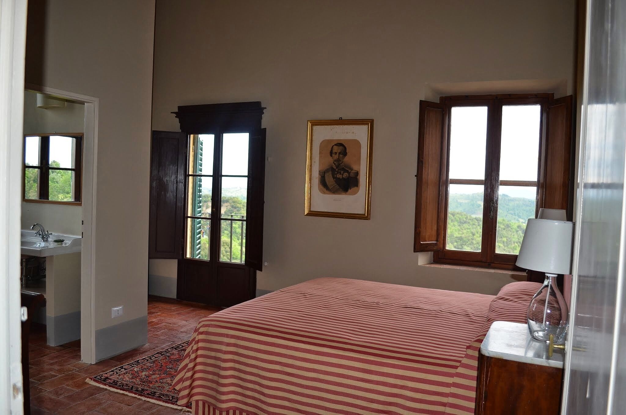 A bedroom with a bed, a window with a view, a framed picture on the wall, and a bedside table with a lamp.