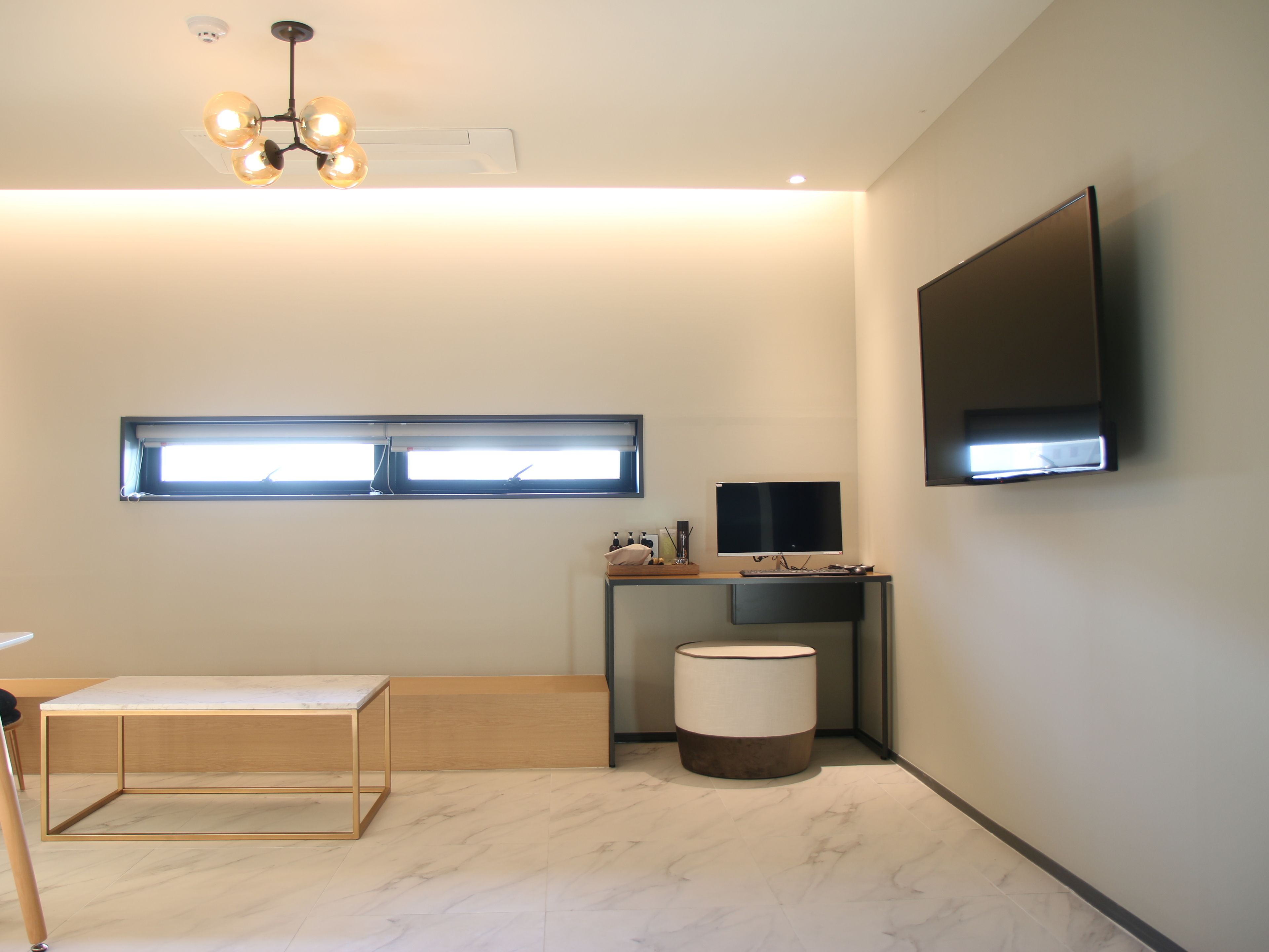 Une chambre moderne et minimaliste, dotée d’un téléviseur à écran plat fixé au mur, d’un petit bureau avec un ordinateur, d’un pouf rond et d’une fenêtre avec des stores.