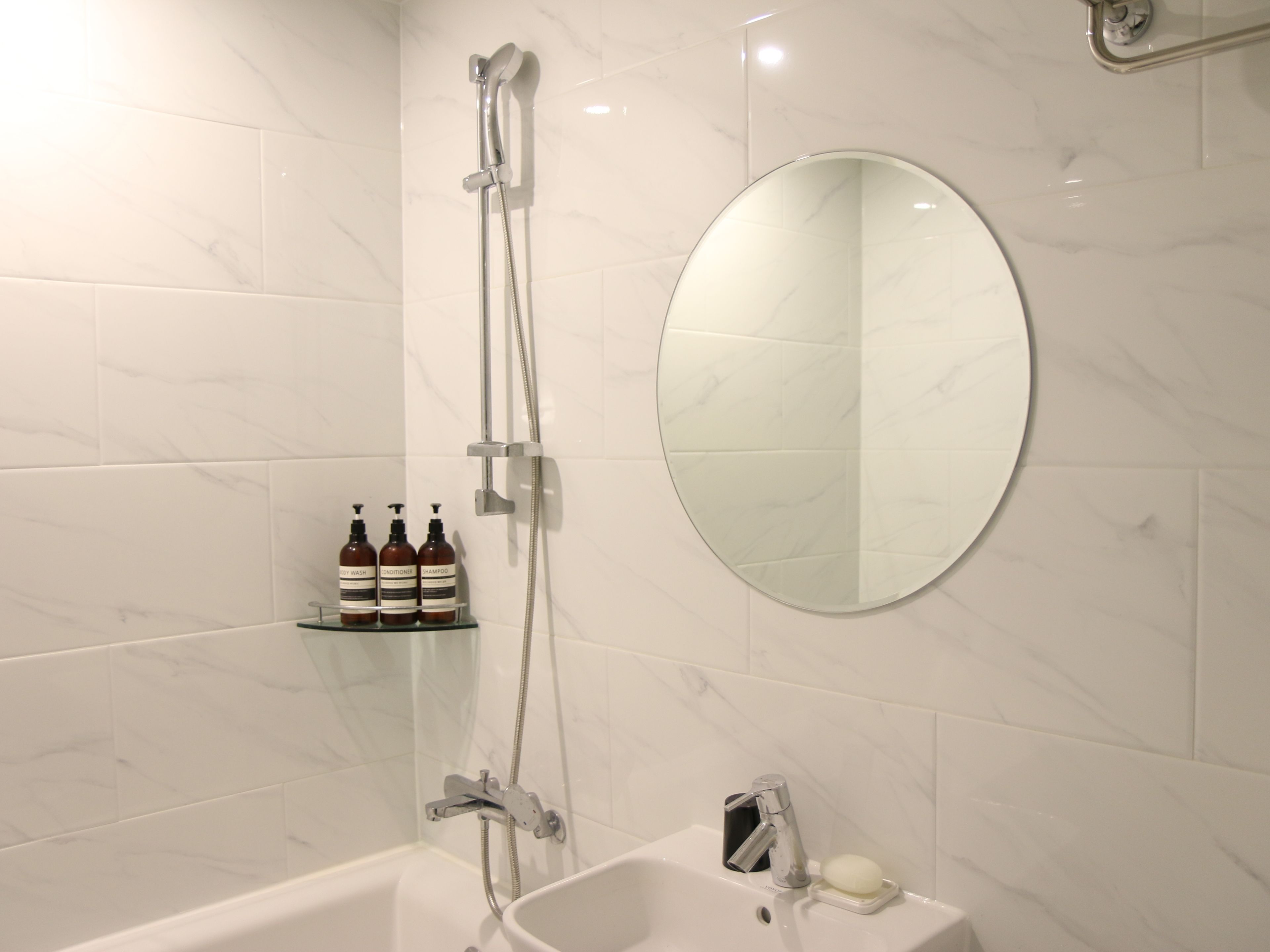 Une salle de bain avec une baignoire, un miroir rond et une étagère sur laquelle sont posées trois bouteilles de shampoing et d’après-shampoing.