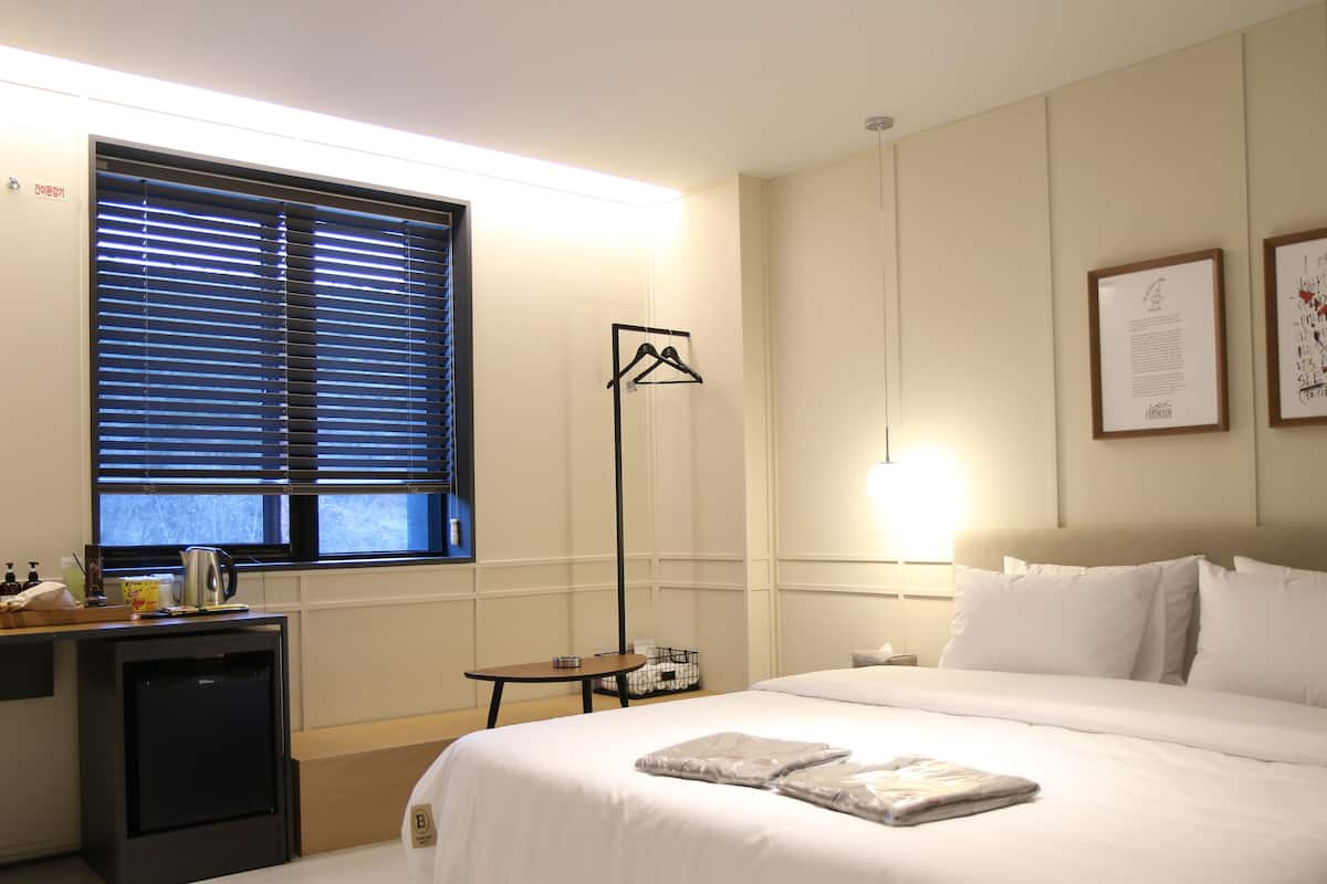 A hotel room with a bed, bedside table, lamp, and a window with blinds.