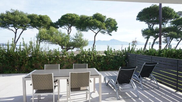 An outdoor patio with a table and chairs, surrounded by greenery and overlooking a body of water with trees and mountains in the distance. - Ground Floor With Stunning Sea Views And Private Terrace, Shared Pool (Pollença)