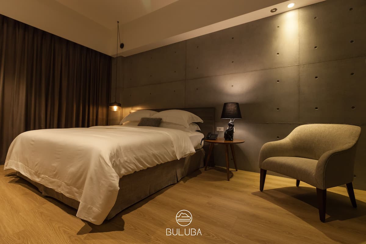 A modern bedroom with a large bed, a chair, a bedside table with a lamp, and a wall with a textured panel.