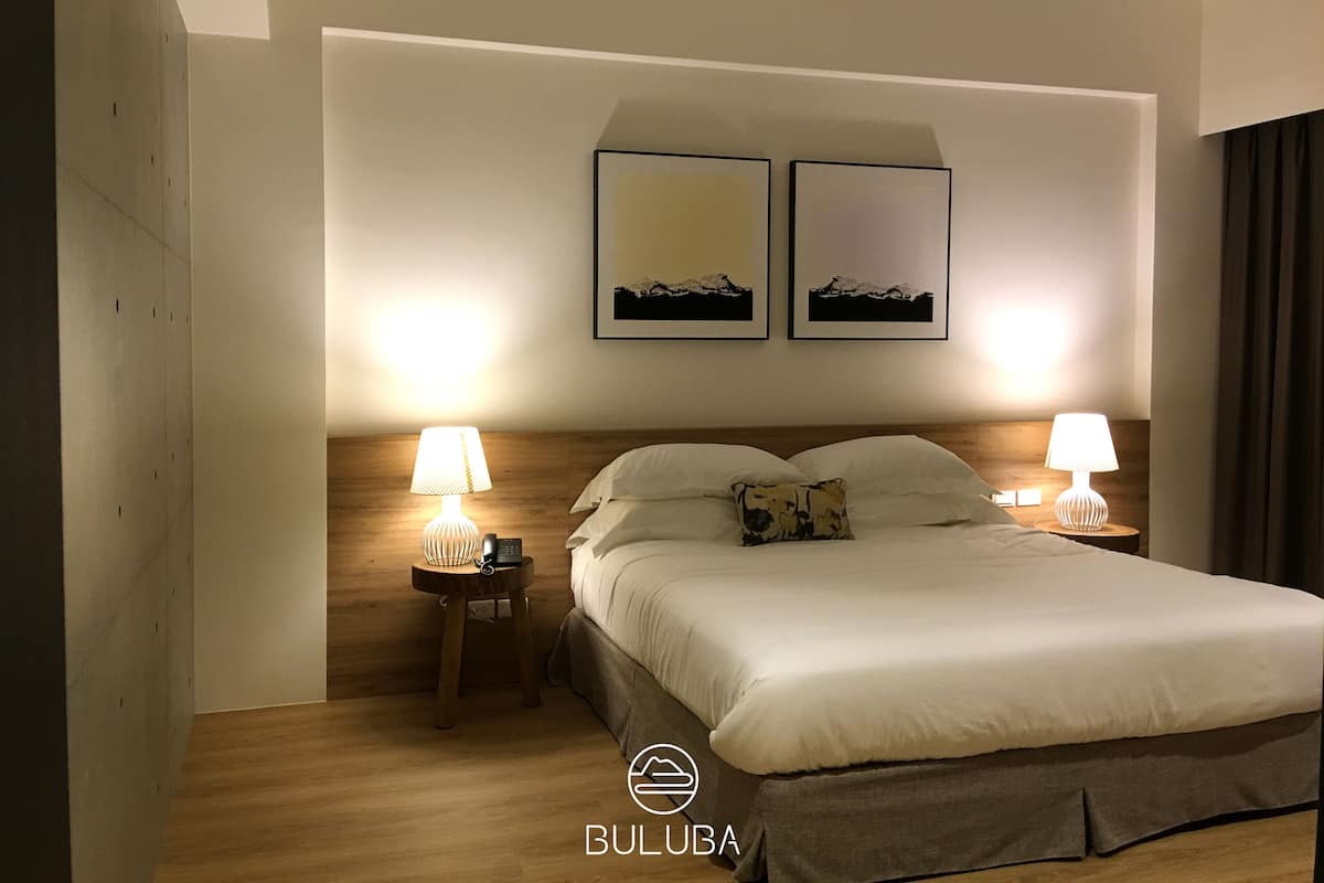 A hotel room with a bed, two bedside lamps, a wooden headboard, and two framed pictures on the wall.