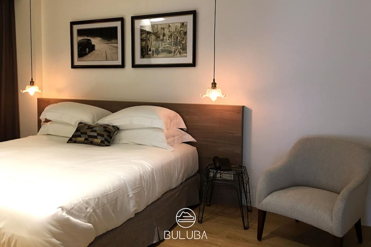 A hotel room with a bed, two framed pictures on the wall, a chair, and a bedside table with a phone.