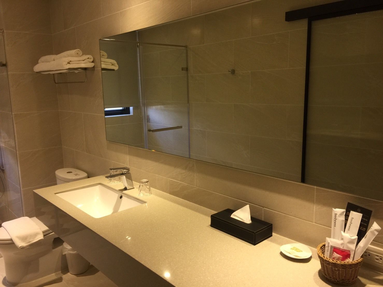 A modern bathroom with a large mirror, a sink, and a toilet. There are towels on a rack and a basket with toiletries.