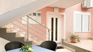 Eine Terrasse mit kariertem Tischlein, Korbstühlen, einem Blumentopf und einer Treppe, die zu einem Innenbereich führt.
