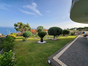 Un jardín bien cuidado con varios árboles y arbustos, un sendero pavimentado y vista al mar.