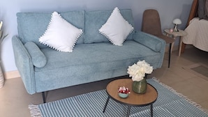Un salon avec un canapé bleu clair, deux coussins blancs ornés de glands, une petite table ronde sur laquelle est posé un vase de fleurs et un tapis à motifs rayés.