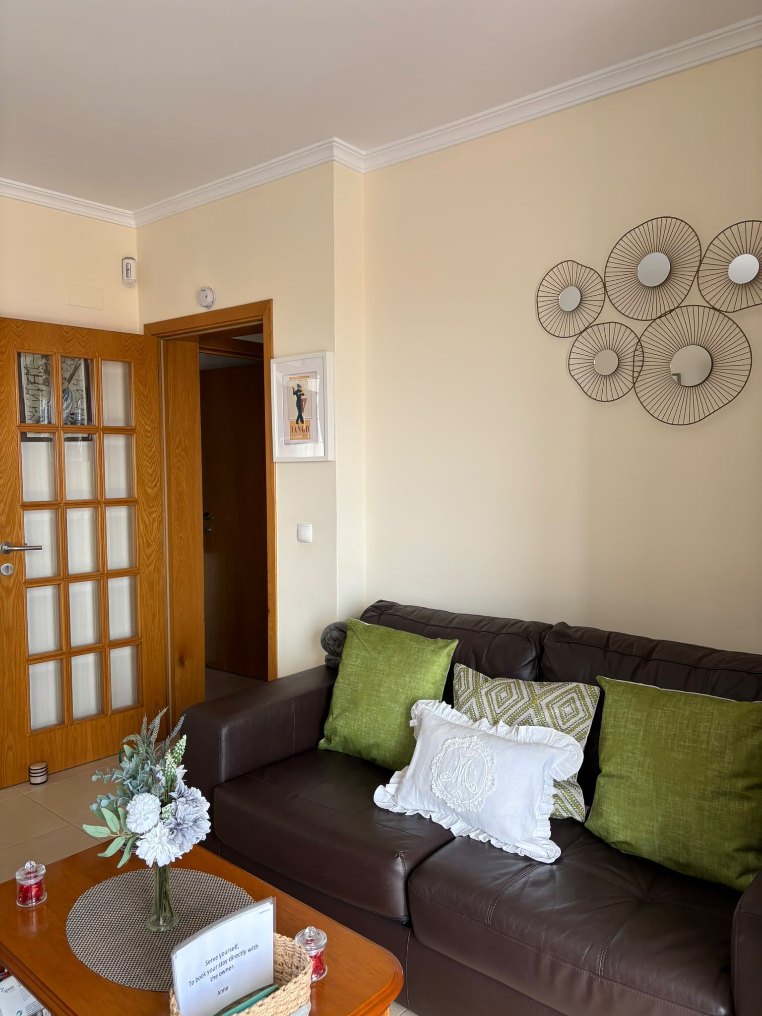 Un salon avec un canapé en cuir marron, des coussins verts et blancs, une table basse en bois ornée d’un vase de fleurs et un élément décoratif fixé au mur.