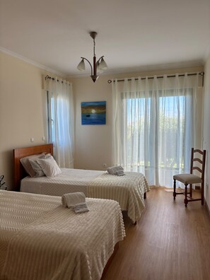 Quarto de hotel com duas camas, uma cadeira de madeira, um quadro e vista para o exterior.
