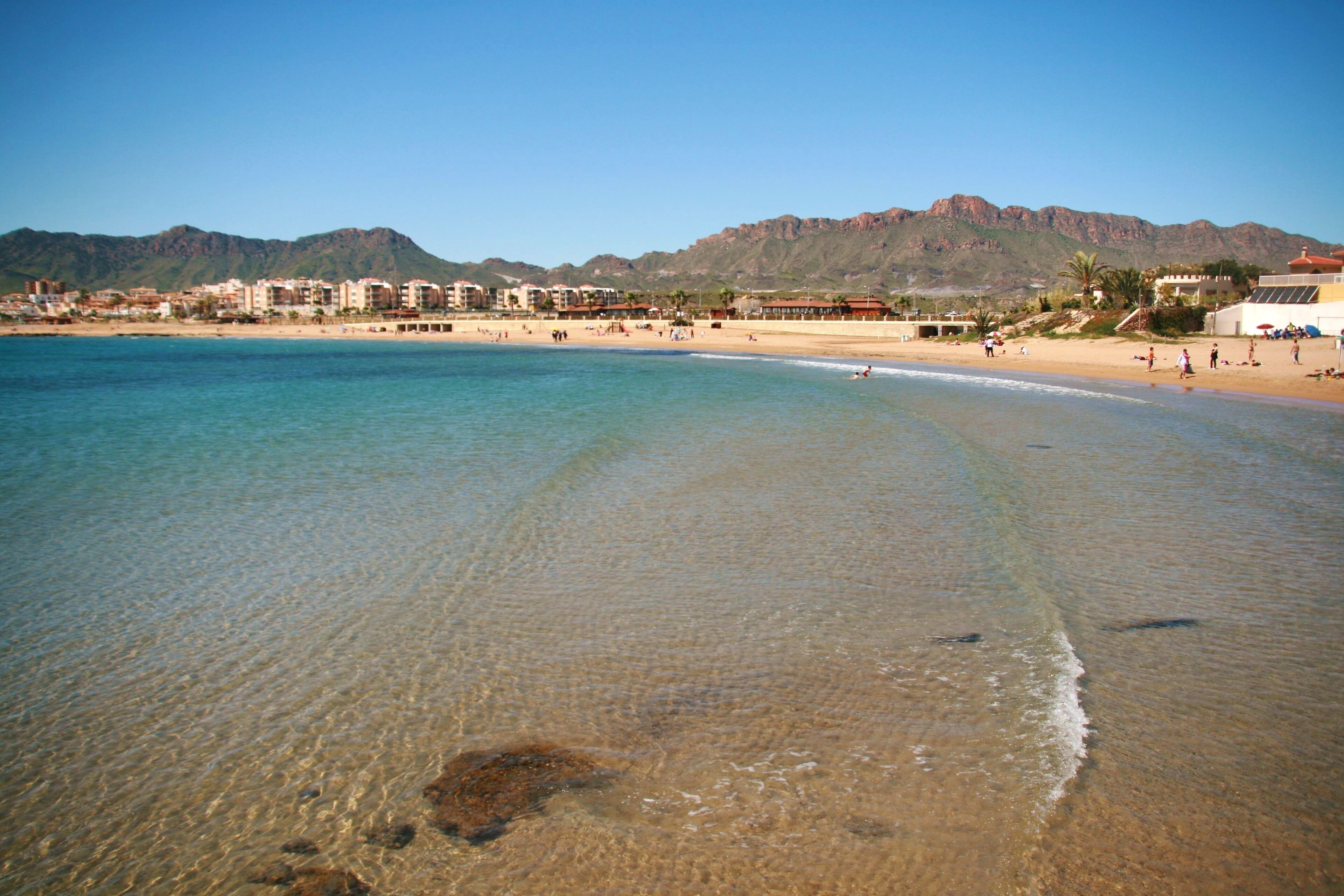 Una spiaggia con acque limpide, una riva sabbiosa e edifici sullo sfondo.