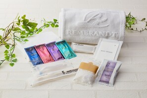 Ein weißes Handtuch mit dem geprägten Schriftzug "REMEMBRANDT", eine Haarbürste, ein Badeschwamm, eine Zahnbürste und eine Packung Lavendel-Badesalz.