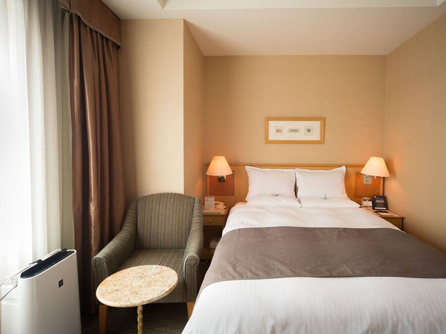 A hotel room with a bed, two bedside lamps, a chair, a small table, and a wall-mounted picture.