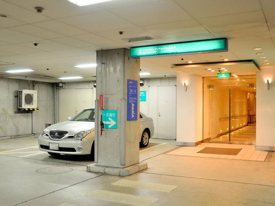 A parking garage with a white car parked near a pillar, a directional sign, and a hallway leading to an elevator.