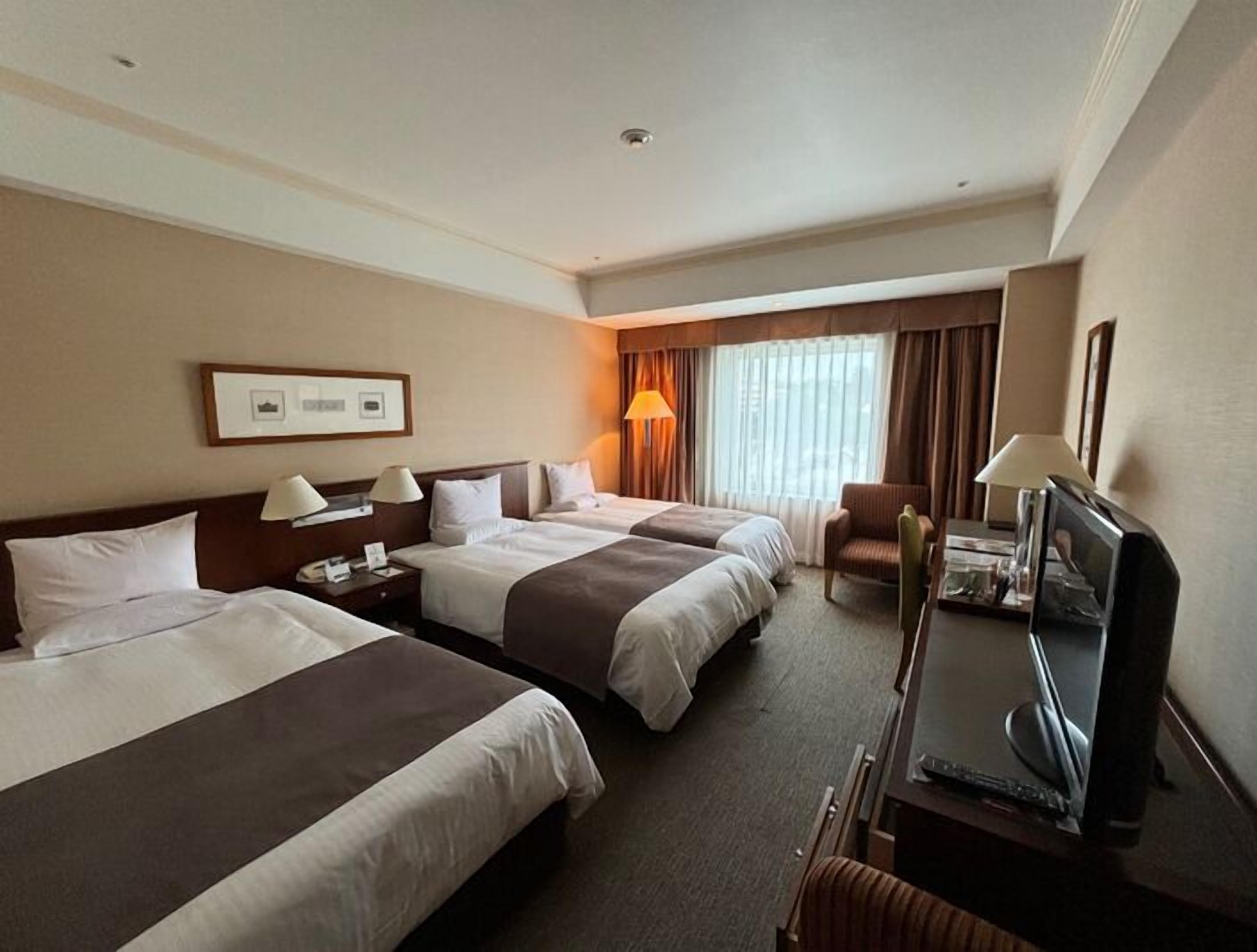 Habitación de hotel con dos camas, un escritorio con televisor, una silla y una ventana con cortinas.