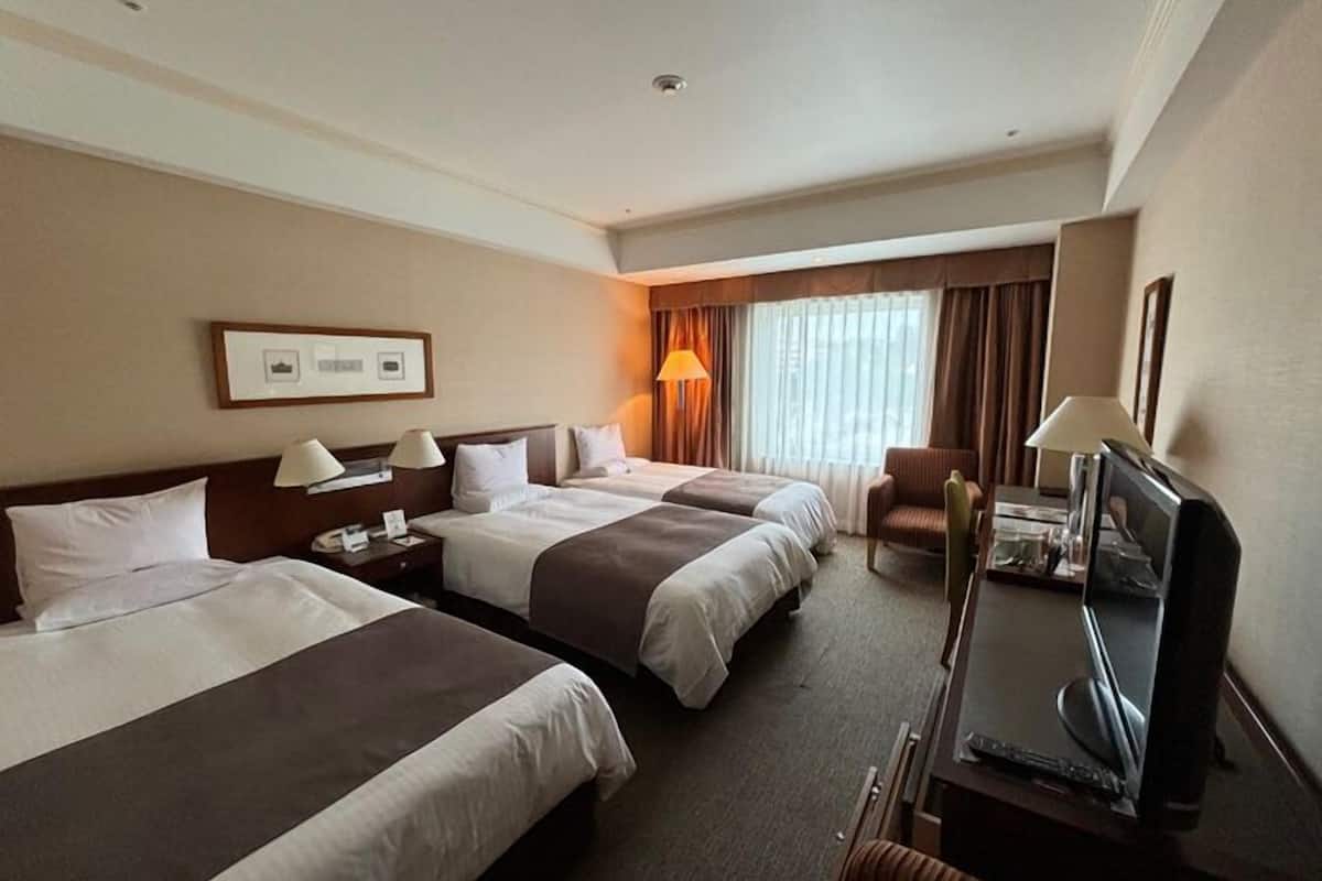 Ein Hotelzimmer mit zwei Betten, einem Schreibtisch mit Fernseher, einem Sessel und einem Fenster mit Vorhängen.