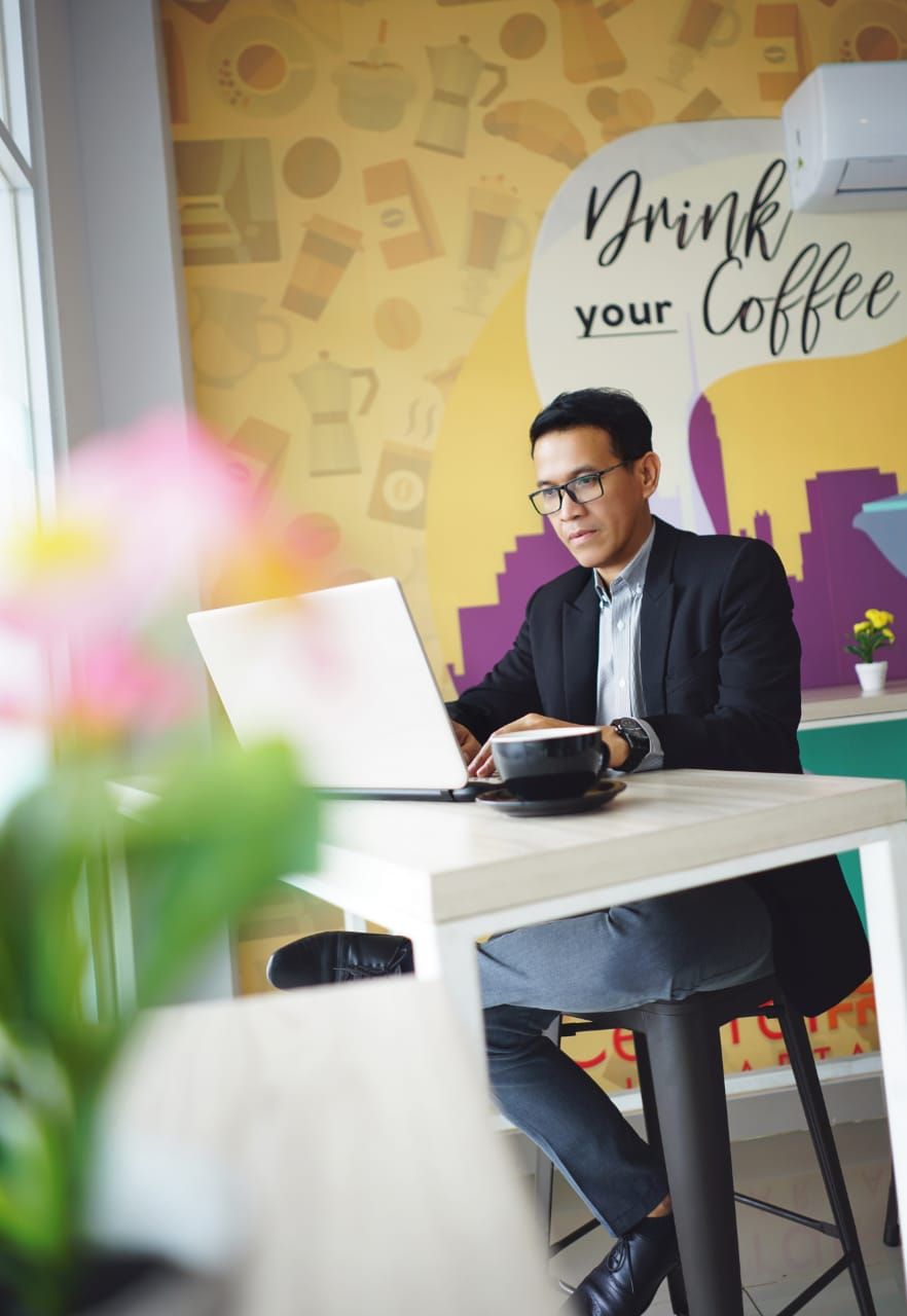 A person sitting at a table with a laptop, in a room with a wall that has a coffee-related design.
