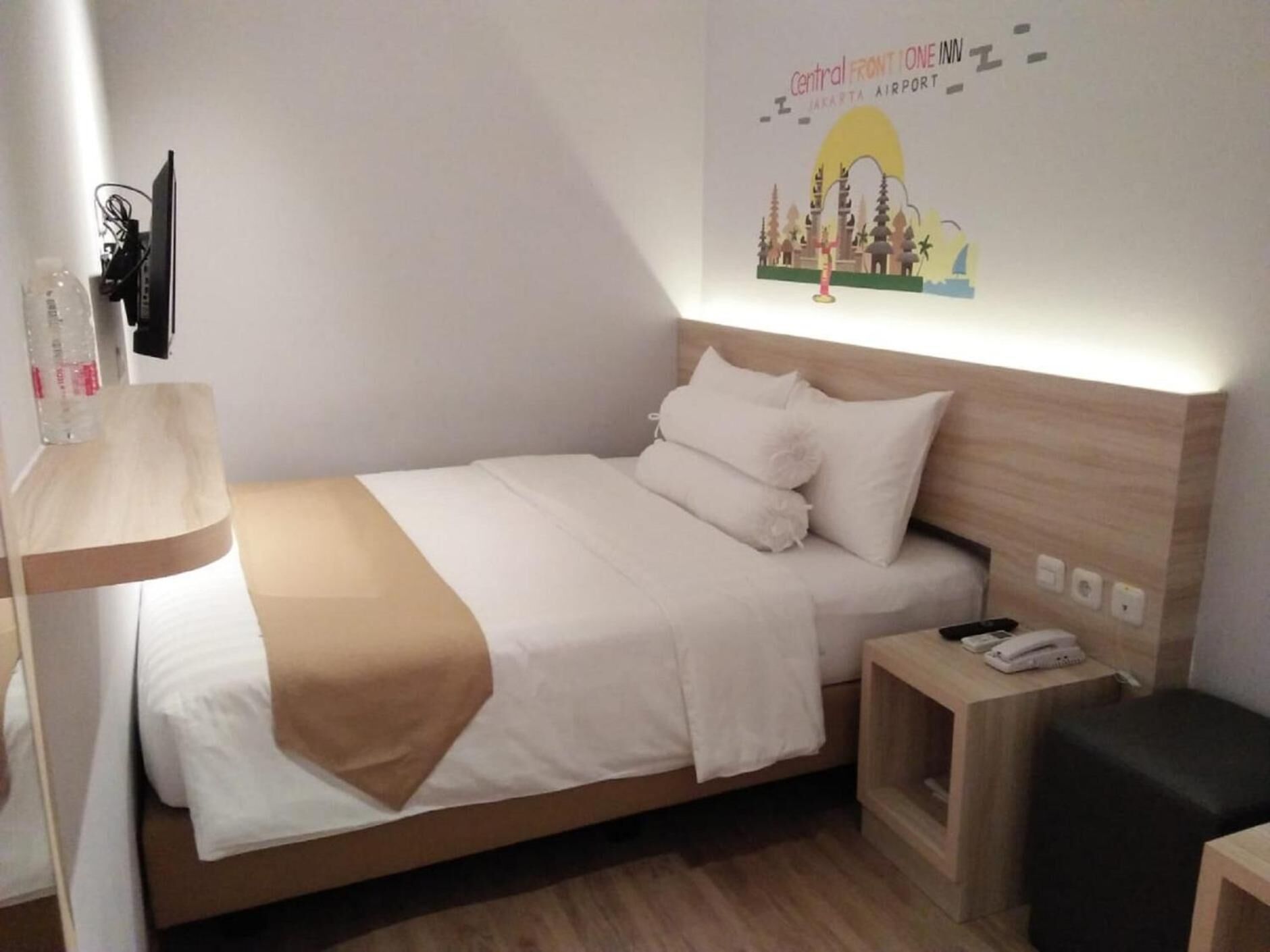 Ein ordentlich bezogenes Bett mit weißer Bettwäsche und einer braunen Tagesdecke, ein Nachttisch mit einem Telefon und eine Stadtlandschaft als Wanddekoration.
