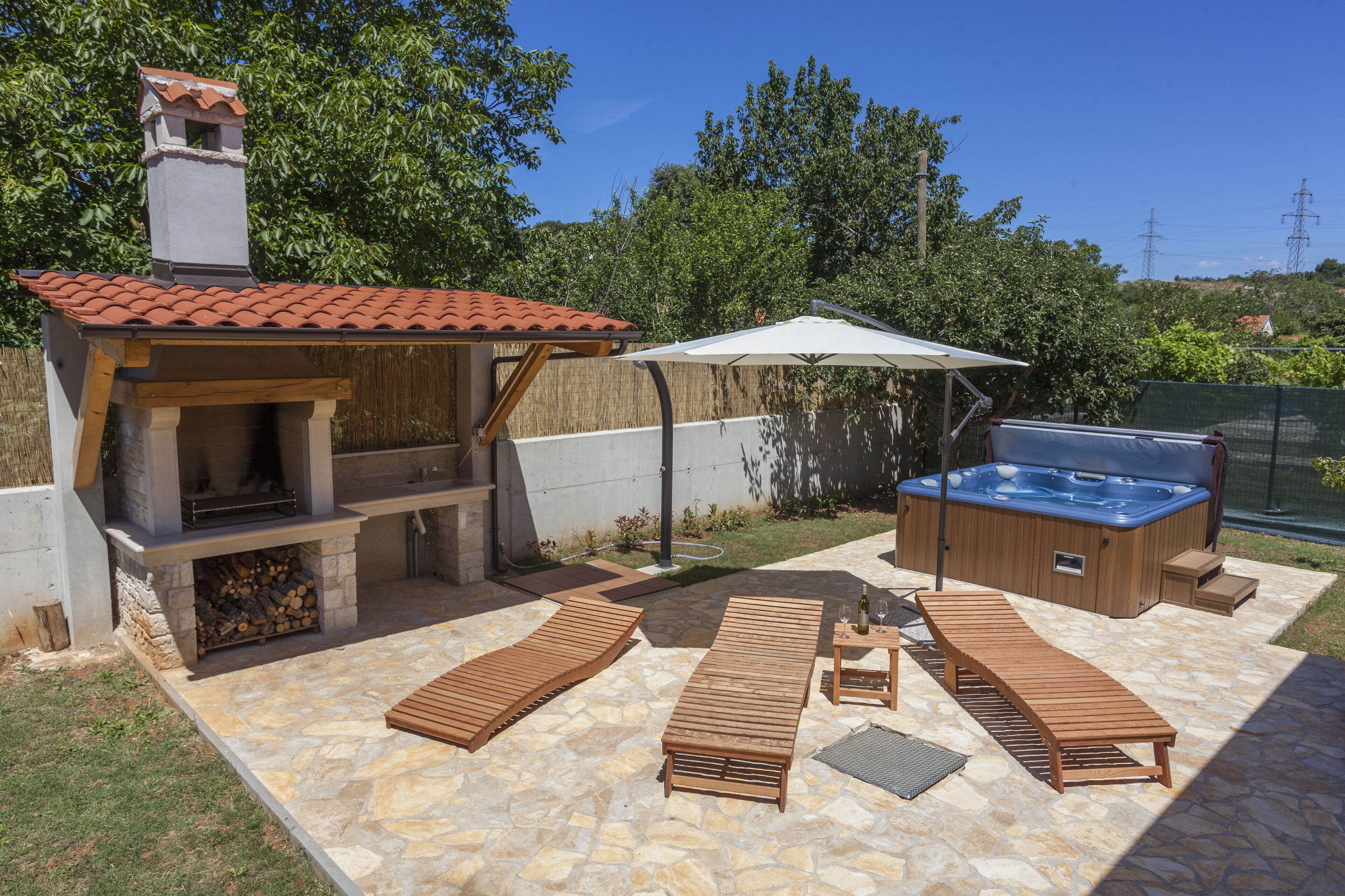 Patio esterno con pavimento in pietra, poltrone da lounge in legno, area barbecue e vasca idromassaggio.