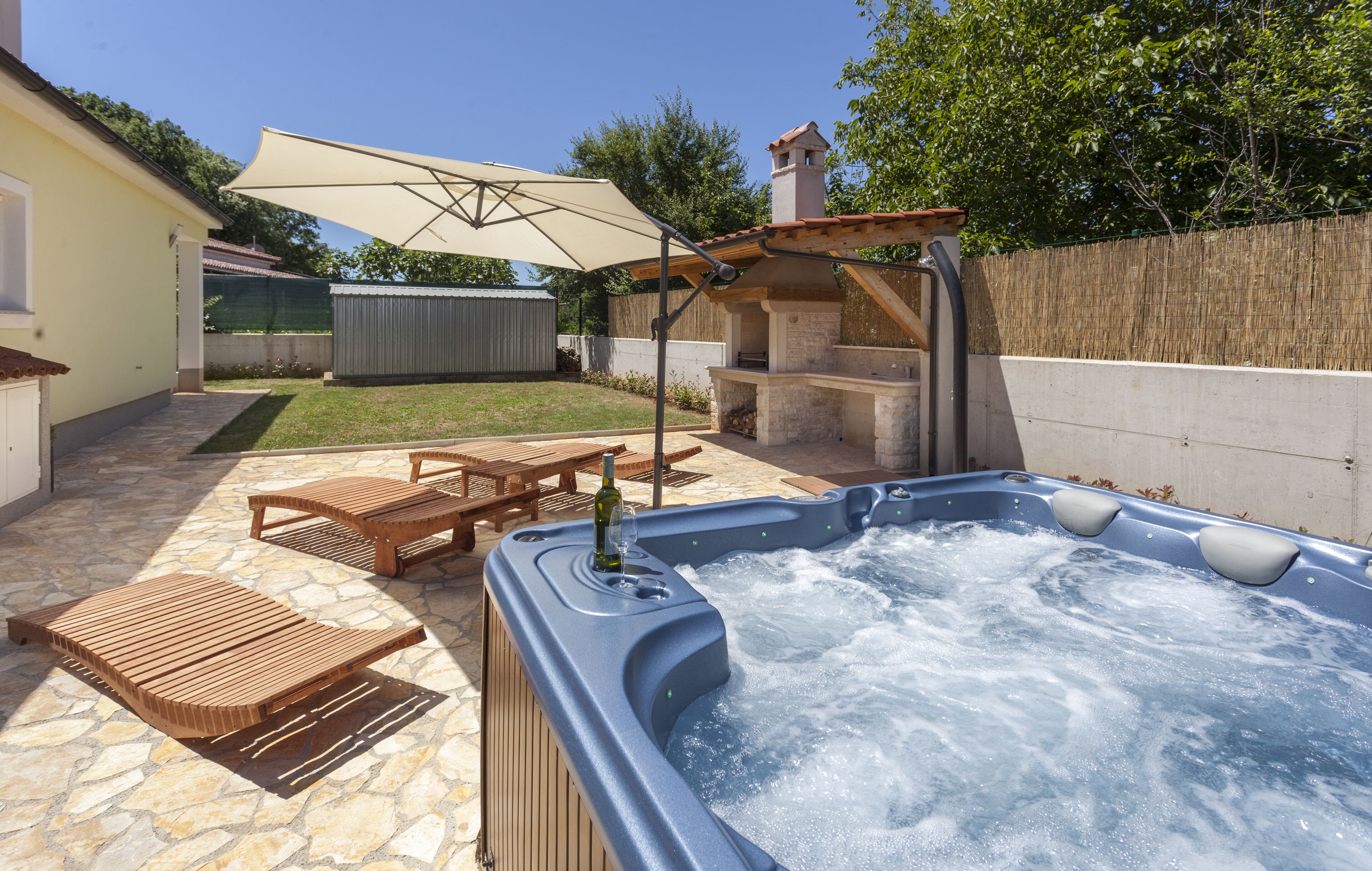 A backyard with a hot tub, wooden lounge chairs, a picnic table, and a barbecue area.