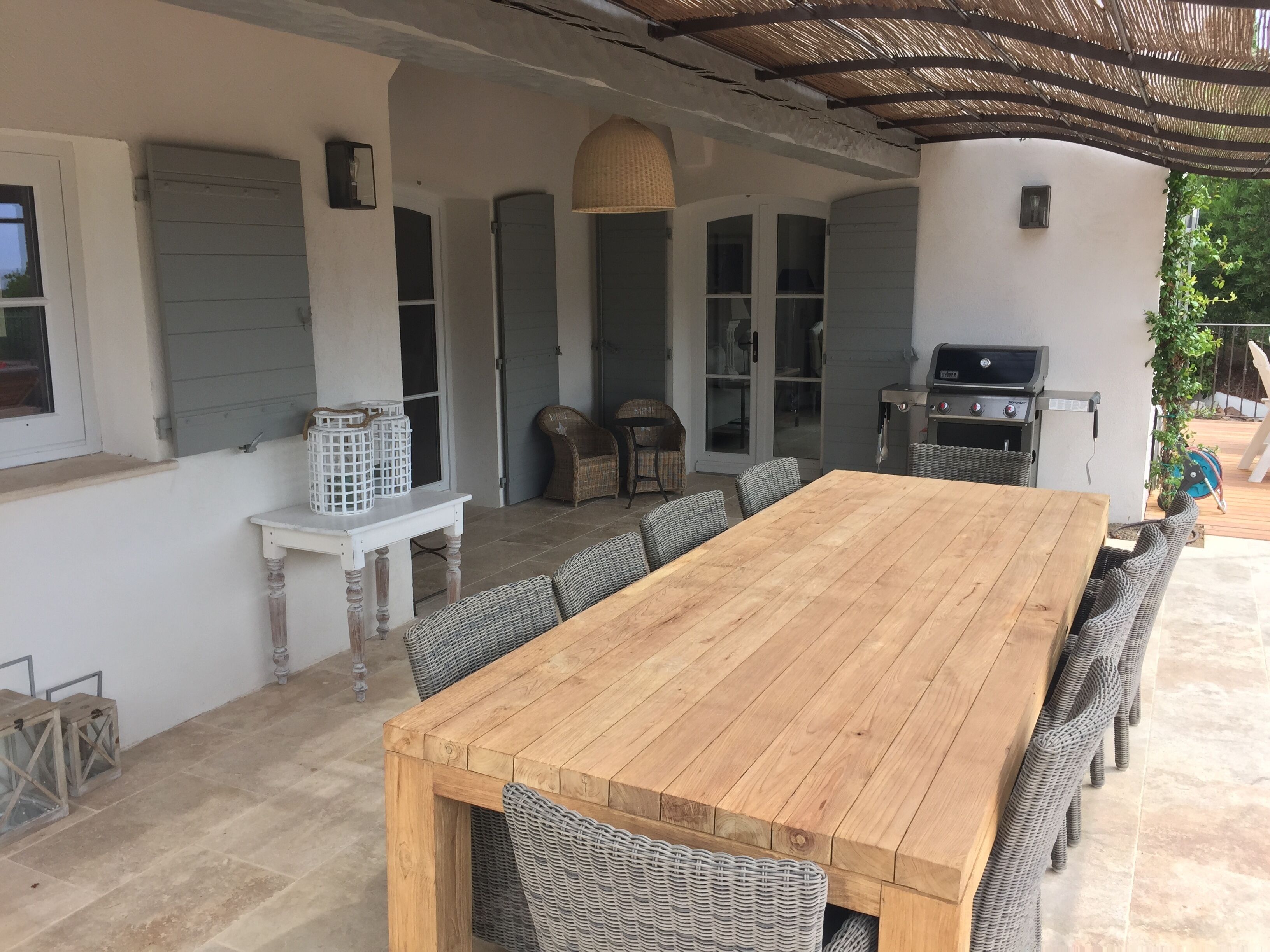 A covered patio area with a wooden table and wicker chairs, a grill, and a basket of laundry.