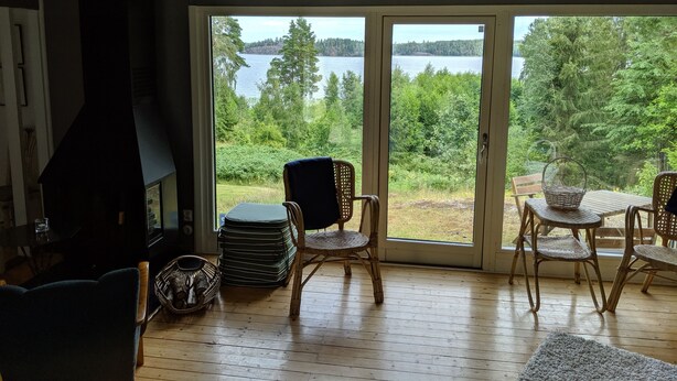 Ein gemütliches Wohnzimmer mit Kamin, Korbsesseln und Blick auf einen See und Wald.