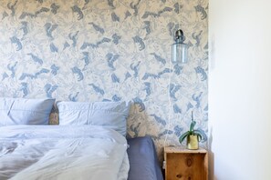 Un dormitorio con papel tapiz estampado, una cama con ropa de cama blanca y azul, una mesita de noche con una planta y una lámpara de pared.