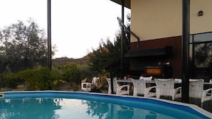 Une piscine entourée de chaises blanches, un espace barbecue et un bâtiment doté de grandes fenêtres.