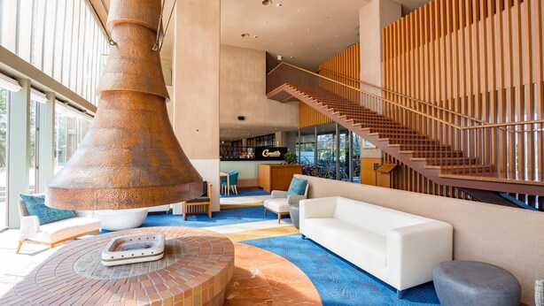 Un vestíbulo amplio con una chimenea cilíndrica grande, una escalera y una zona de estar con un sofá blanco y una mesa redonda.