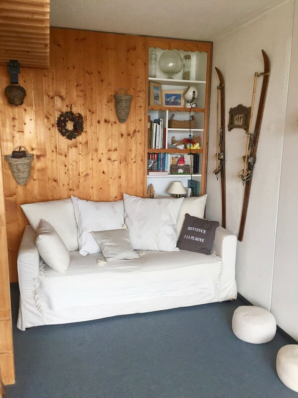 A white sofa with a 'Bienvenue A La Plagne' pillow, a bookshelf with books and decorative items, and skis leaning against the wall. - Charming 2 room apartment - view of Mont-Blanc - at the foot of the slopes in Aime 2000 (Plagne Aime 2000)